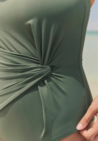Olivgrünes Badeanzug mit einem verdrehten Knoten-Detail an der Taille, gefertigt aus glattem, elastischem Stoff mit schmaler Silhouette.