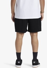 Persona che indossa pantaloni sportivi in rete neri, calze nere, scarpe da ginnastica nere della marca DC e una maglietta bianca, vista da dietro.