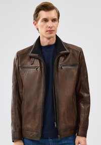 Veste en cuir marron avec une texture lisse, fermeture �éclair, deux poches avant et un col côtelé. Portée sur un pull bleu marine.