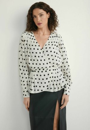 Kobieta o kręconych włosach, ubrana w białą bluzkę w kropki z długim rękawem oraz czarną spódnicę z rozcięciem z przodu, stojąca na jednolitym tle.