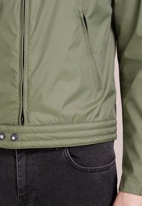 Veste isolante verte avec une fermeture éclair à l'avant et des poches latérales, présentant une texture lisse et une ceinture côtelée. Un jean noir est visible en dessous.