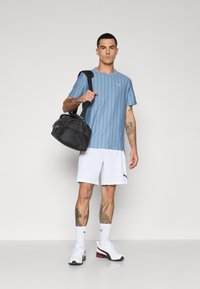 Lichtblauw gestreept sportshirt, witte shorts, zwarte sporttas en witte schoenen met rode accenten, staand tegen een effen achtergrond.