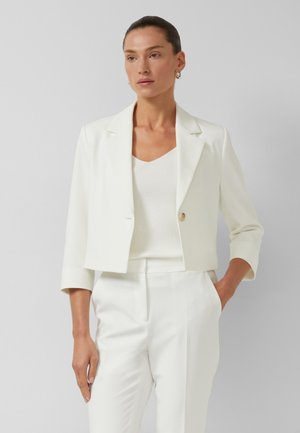 Vrouw met haar naar achter gebonden, draagt een witte cropped blazer, witte top en witte hoge taillebroek, staand met één hand in haar zak.