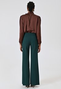 Blouse en satin marron avec des manches bouffantes et un col haut, associée à un pantalon large taille haute vert foncé, mettant en valeur une silhouette ajustée.