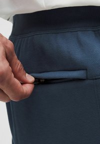 Marineblauwe sportbroek met een gladde textuur, voorzien van een verborgen ritszak aan de zijkant en een elastische tailleband voor comfort.