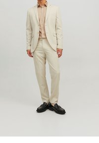 Lichtbeige pak met een nauwsluitende pasvorm, een colbert met één rij knopen en bijpassende broek. Geaccentueerd door een lichtbeige overhemd met knopen en zwarte schoenen.