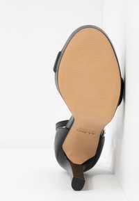 Chaussure à talons hauts noire avec une tige en cuir lisse et une semelle extérieure en caoutchouc beige clair dotée de rainures horizontales pour l'adhérence.