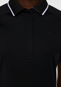 Svart pikétröja med en texturerad krage som har en vit rand, knappstängning och en liten svart logotyp på vänster sida.