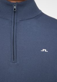 Tummansininen vetoketjullinen neulepusero, jossa on korkea ribattu kaulus, teksturoitu kangas ja pieni valkoinen logo vasemmalla puolella.