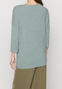 Vala mintunvihreä, ylisuuri pitkähiha-paita pehmeästä kankaasta, laajalla kaula-aukolla ja pudotetuilla harteilla, yhdistettynä neutraaleihin leveälahkeisiin housuihin.