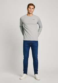 Grijze langemouwen shirt met een ronde hals, gecombineerd met donkerblauwe slim-fit jeans en witte sneakers. Het heeft een klein logo op de borst.