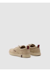 Zapatos de ante beige con un perfil bajo, punta redonda y una lengüeta en el talón a rayas negro-rojo en contraste. Presenta cordones tonales y una suela texturizada.