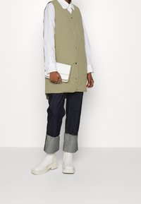 Gequilted olijfgroene vest met knopen, gedragen over een witte blouse met kraag. Donkerdenim jeans met een rafelige zoom en witte hoge enkellaarzen. Houdt een witte clutch vast.