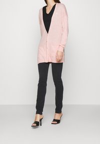 Světle růžový dlouhý kardigan s výstřihem do V, zapínáním na knoflíky a dvěma kapsami, nošený přes černý top s výstřihem do V, doplněný tmavými džínami a černými podpatky.