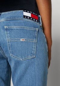 Osoba nosząca jasnoniebieskie dżinsy z denimu i czarną bluzkę, pokazująca naszywkę z logo Tommy Jeans obok pasa oraz małe logo na tylnej kieszeni.