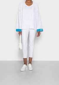 Bílý lehký cardigan s otevřeným předním dílem, který má texturovaný vzor a tyrkysové manžety; spárovaný s bílými přiléhavými kalhotami a teniskami.