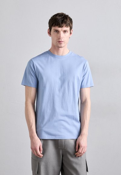 Tricou din bumbac de culoare albastru deschis, cu decolteu clasic și mâneci scurte, asortat cu pantaloni cargo gri, stând în fața unui fundal neutru.