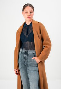 Femme portant un long manteau beige sur un haut en dentelle marine et un jean gris taille haute, se tenant avec une main dans sa poche contre un fond blanc.