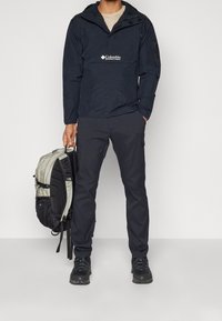 Man som bär en svart Columbia vindjacka, svarta byxor och vandringsskor, håller en grå och svart ryggsäck i en hand.