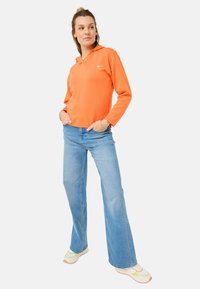Oranje hoodie met een voorzak en wit logo, gecombineerd met lichtblauwe flared jeans en meerkleurige sportschoenen.