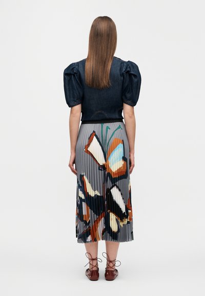 Haut en denim à manches bouffantes associé à une jupe plissée imprimée de papillons multicolores. Le mannequin porte des chaussures marron à lacets.