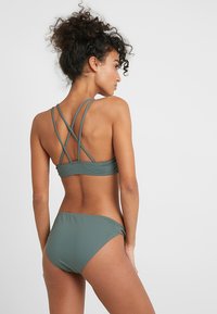 Zelený bikiny set s prekríženým dizajnom na chrbte. Vyrobený z hladkej tkaniny, obsahuje dvojité ramienka a nohavičky v hipster štýle so bočnými akcentmi.