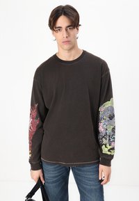 Tmavě hnědé tričko s dlouhým rukávem, které má barevné grafické potisky na obou rukávech, uvolněný střih a kulatý výstřih. Kombinováno s modrými džíny.