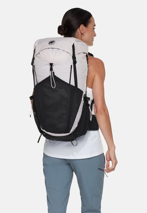 Kvinde iført en stor lysegrå og sort vandrerygsæk med et logo på øverste front, klædt i hvid top uden ærmer og blå bukser.