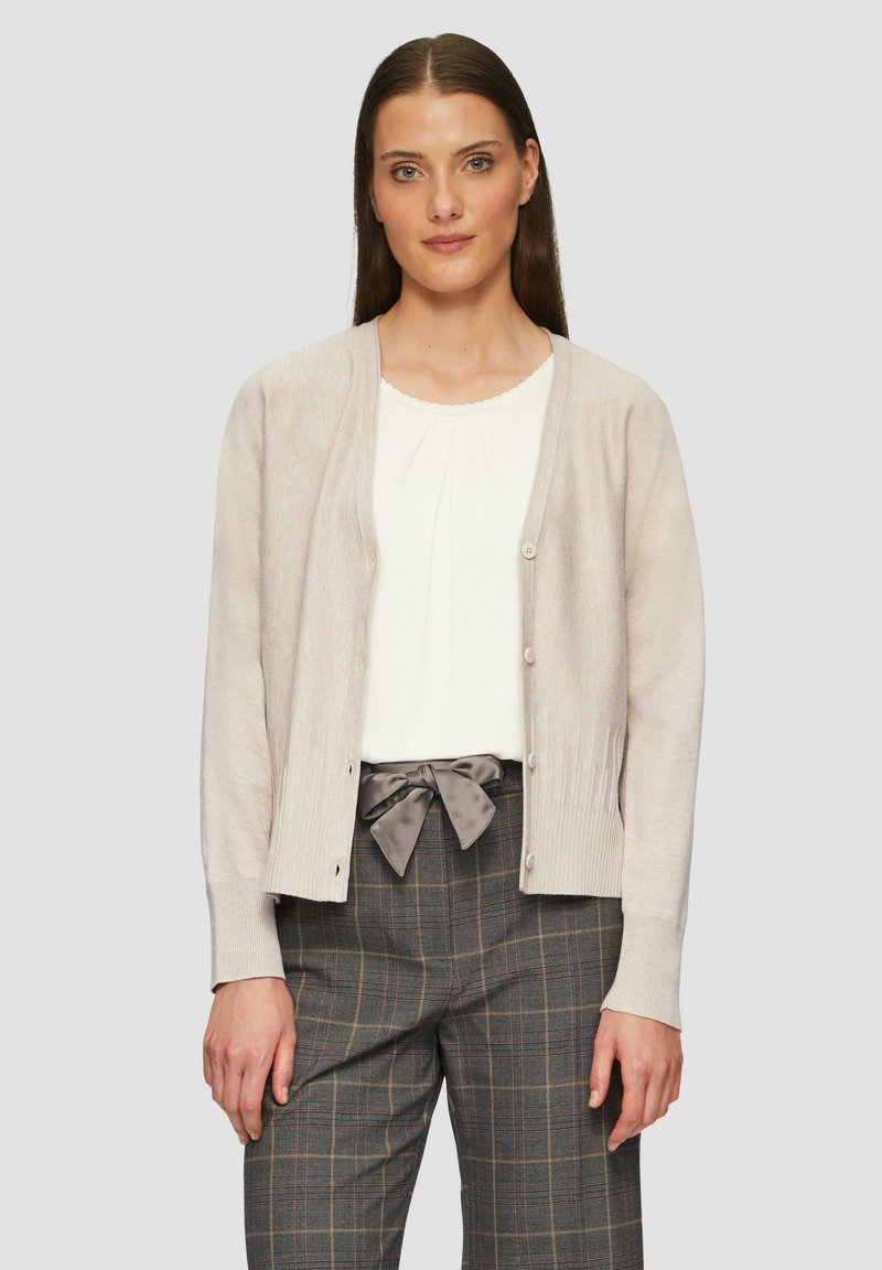 Cárdigan de color beige claro con puños acanalados, botones en la parte delantera, usado sobre una blusa blanca y pantalones de cuadros grises con un acento de lazo.