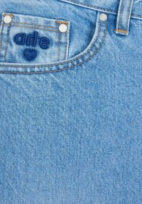 Jeans di denim azzurro chiaro con una tasca frontale che presenta un'etichetta ricamata "arte" e due rivetti metallici. Le cuciture sono di un colore arancione a contrasto.