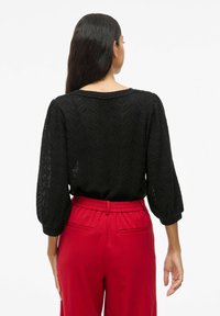 Blouse noire texturée avec un motif en chevrons et des manches bouffantes, associée à un pantalon rouge à taille élastique avec poches arrière.
