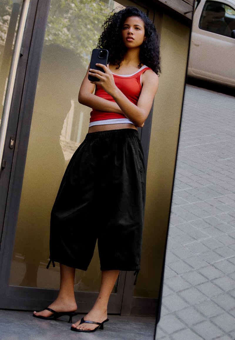 Jeune femme aux cheveux bouclés portant un débardeur rouge et un pantalon noir large prend un selfie devant une porte vitrée réfléchissante à l’extérieur.