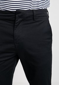 Pantaloni in tessuto nero con una texture liscia, caratterizzati da un taglio classico e passanti per cintura. La vita è leggermente sopra i fianchi, sagomati in modo preciso.
