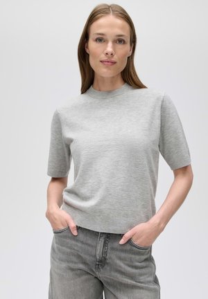 Femme aux cheveux lisses châtain clair porte un top en maille gris clair à manches courtes et un jean gris délavé, les mains dans les poches, sur un fond uni.