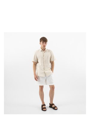 Junger Mann steht, trägt ein beige kurzärmeliges Hemd mit Knöpfen, weiße Shorts und dunkelbraune Sandalen vor weißem Hintergrund.