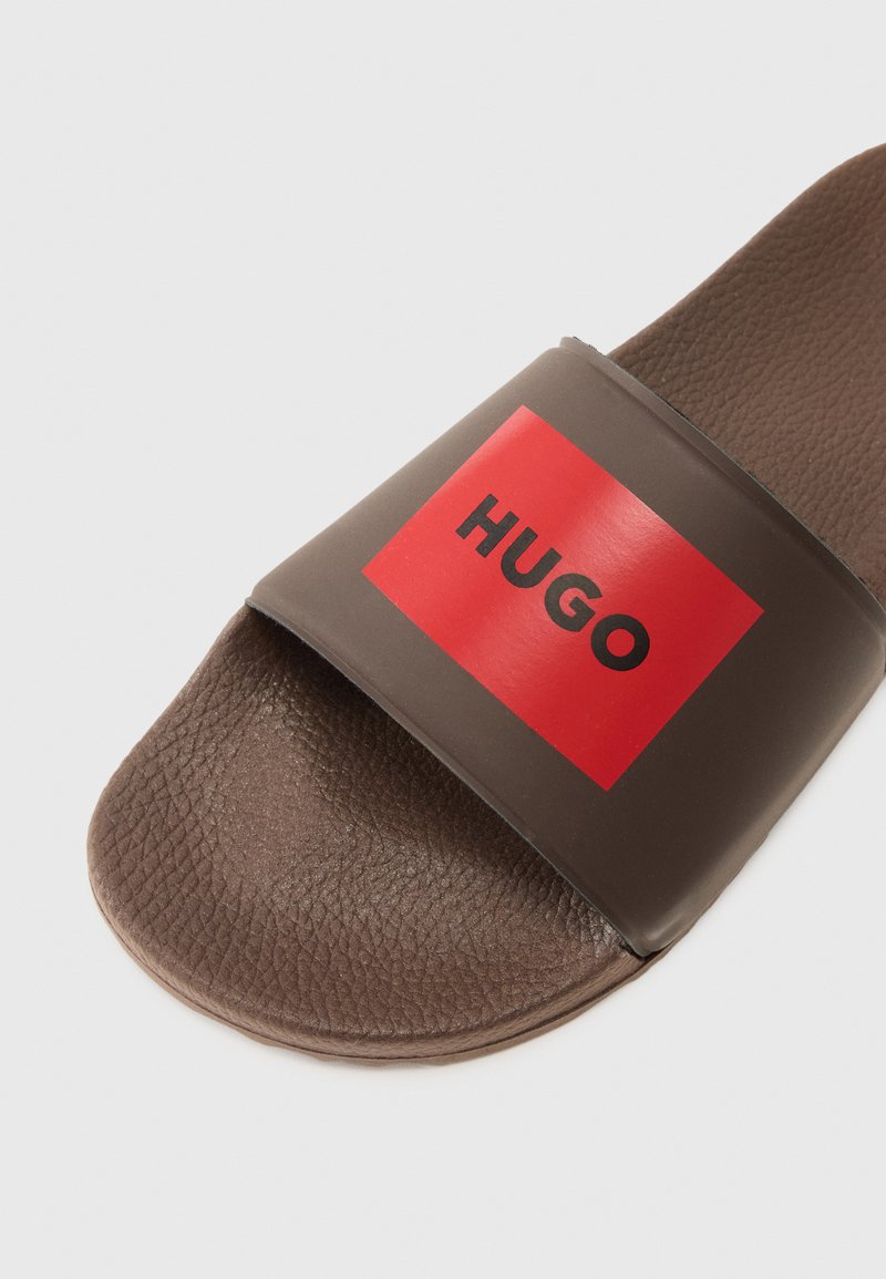 Braune Rutschsandale mit einem breiten Riemen, der ein rotes Rechteck und schwarzen "HUGO"-Text zeigt, strukturiertem Fußbett und glatter Sohle.