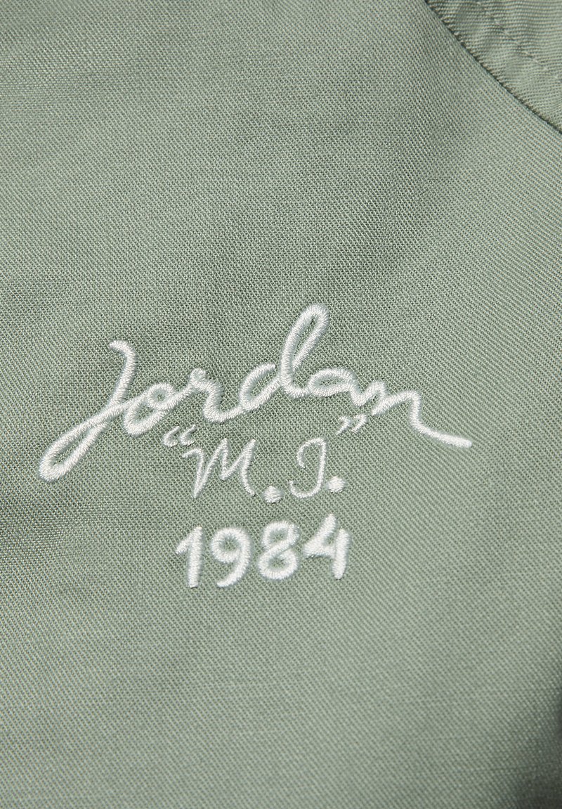 Zielona tkanina z białym haftowanym napisem: "Jordan" w kursywie, "M.I." i "1984" w prostszym kroju, na teksturowanym tle.
