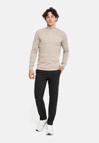 Beige Strickpullover mit hohem Kragen und Frontreißverschluss. Kombiniert mit schwarzen Hosen und weißen Sneakers, was ein lässiges Outfit ergibt.