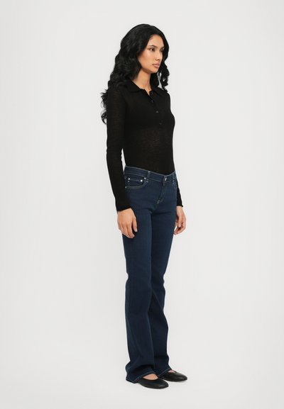 Top negro de manga larga con cuello y botones, combinado con jeans acampanados de color azul oscuro. La modelo se encuentra de lado, mostrando el ajuste y el diseño del atuendo.