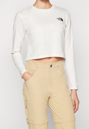 Osoba ubrana w biały krótki top z długim rękawem z logo The North Face oraz beżowe spodnie rozpinane z kieszeniami, z jedną ręką w kieszeni.