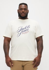 Kremasti T-shirt od pamuka s kratkim rukavima i okruglim izrezom. Sadrži logotip u tamnoplavoj i crvenoj boji na prsima. Bez ukrasa.