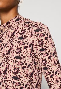 Close-up van een persoon die een beige top draagt met zwart en donkerrode bloemenpatronen en een gouden oorbel in de vorm van een ring.