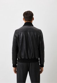 Mężczyzna stojący tyłem, ubrany w czarną skórzaną kurtkę z prążkowanymi mankietami i pasem, na jednolitym jasnym tle.