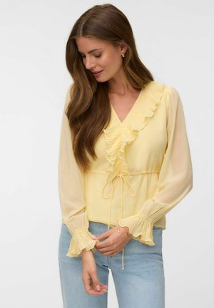 Femme aux longs cheveux bruns portant un chemisier jaune pâle à volants avec des manches transparentes et un jean bleu clair, regardant vers le bas et touchant ses mains.