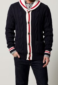 Marinblå kabelstickad cardigan med sjalkrage, vita och röda randiga detaljer, träknappar och två framfickor.