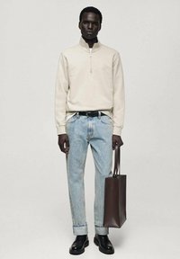 Hanorac crem cu fermoar, blugi de denim cu spălare deschisă, curea neagră și botine masive negre; ținând o geantă tote de culoare maro închis.