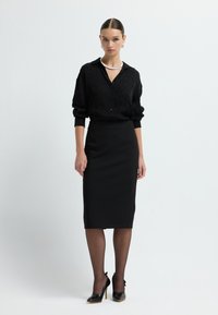 Schwarze Bluse mit Leopardenmuster, schwarzer Bleistiftrock, durchsichtige schwarze Strumpfhosen und schwarze High-Heels mit Schleifen-Detail. Einfache Halskette.