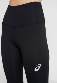 Czarne legginsy sportowe z wysokim stanem, z tonalnym wykończeniem i białym logo na dolnej części nogawki. Gładki, elastyczny materiał.