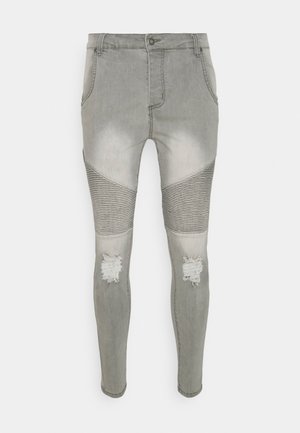 Lysegrå slim-fit jeans med ribbet paneler over knæene og slidte lapper på begge knæ mod en ensfarvet baggrund.