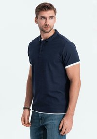 Námořnická modrá polo košile s tříknoflíkovou légou, krátkými rukávy s bílým lemem a klasickým střihem, v kombinaci s modrými džínami.